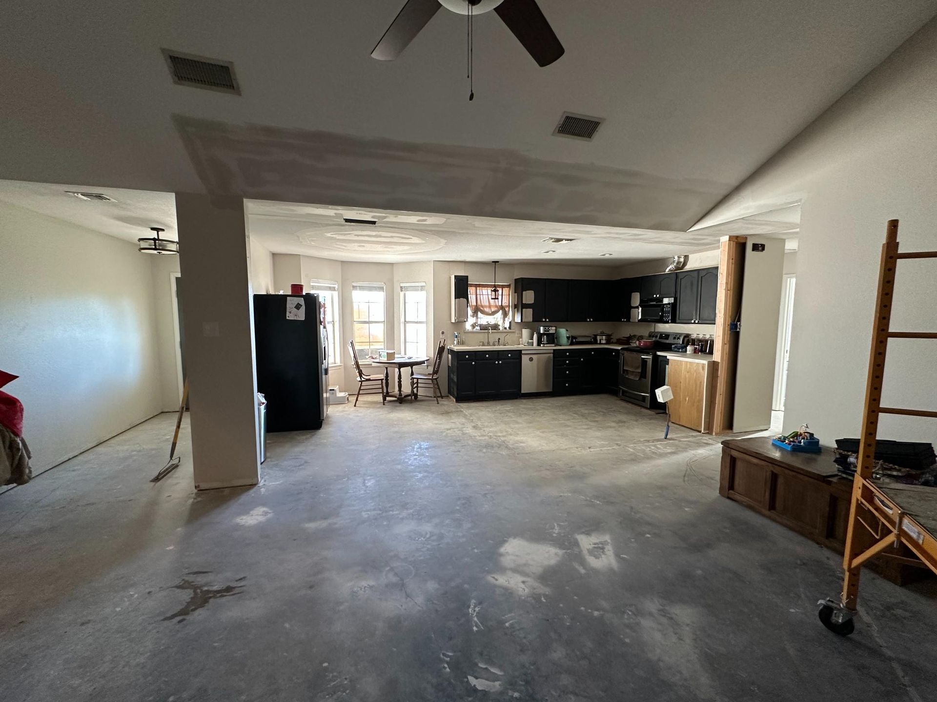 Open-concept kitchen and living area renovation; dark cabinets, unfinished walls, concrete floor, ceiling fans, and ladder.