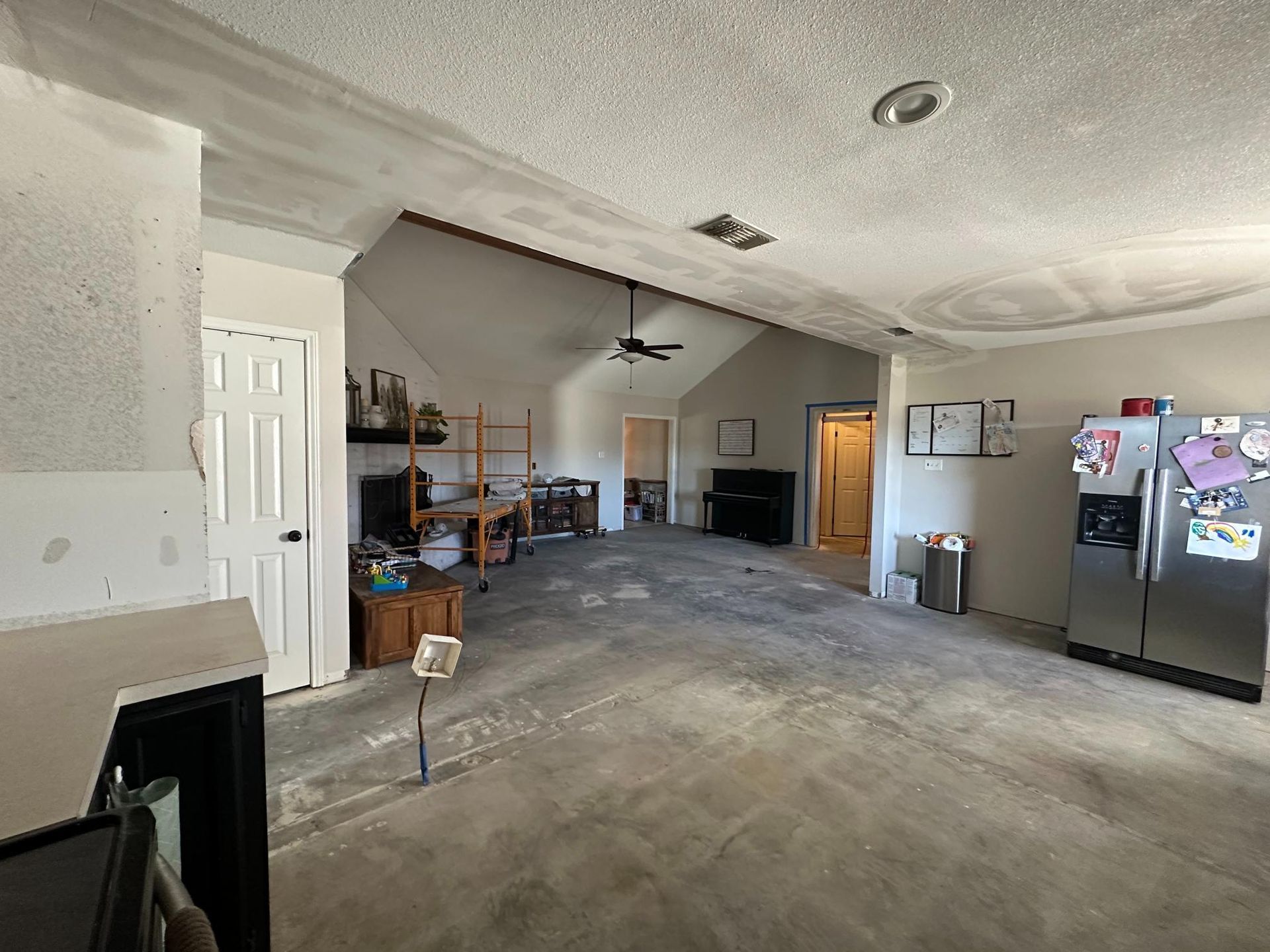 Interior view of a garage with concrete floor, high ceiling. Refrigerator, door, and work area visible.