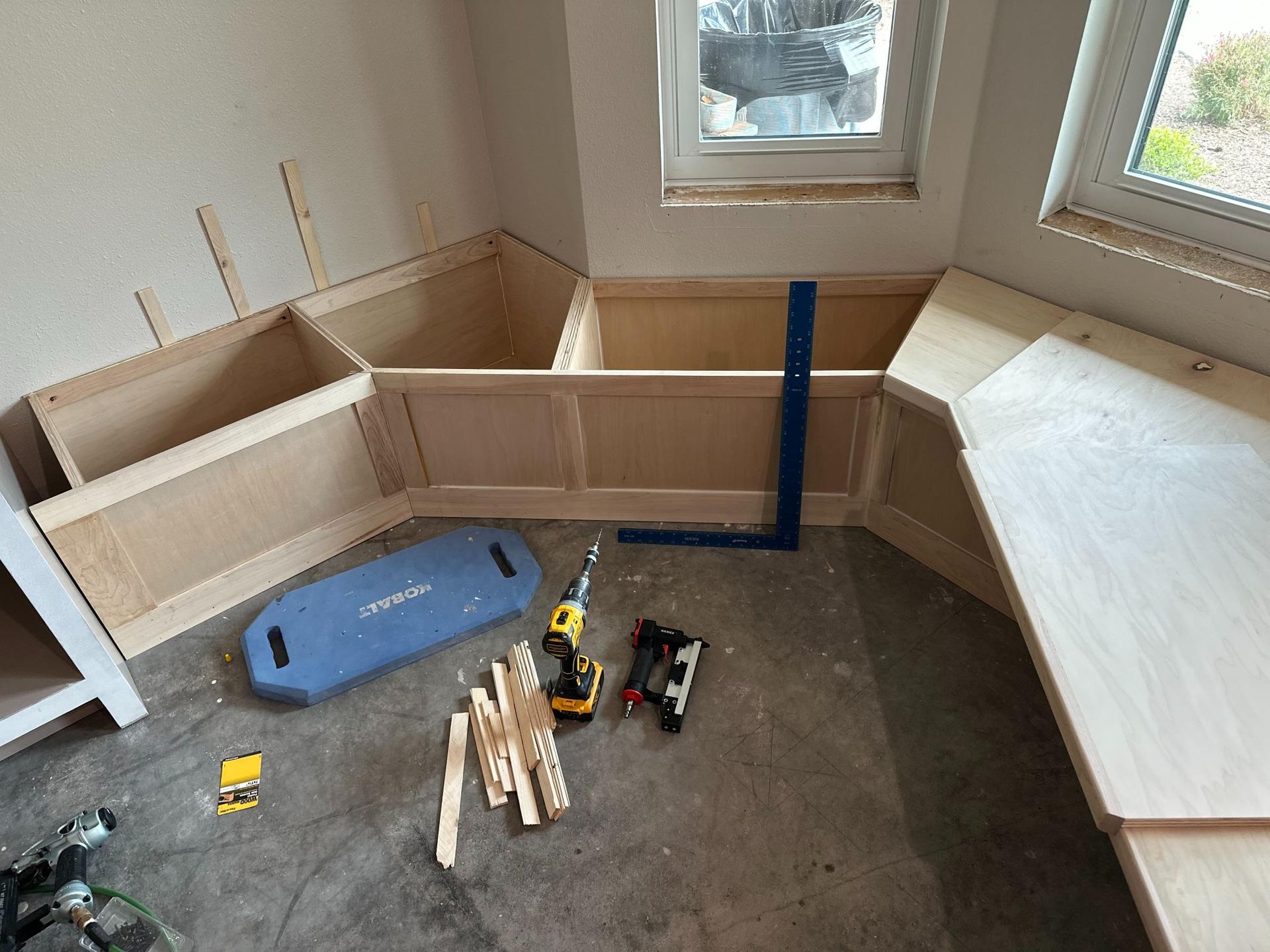 Building a wooden bench seat in a corner near a window. Tools and materials are scattered on the floor.