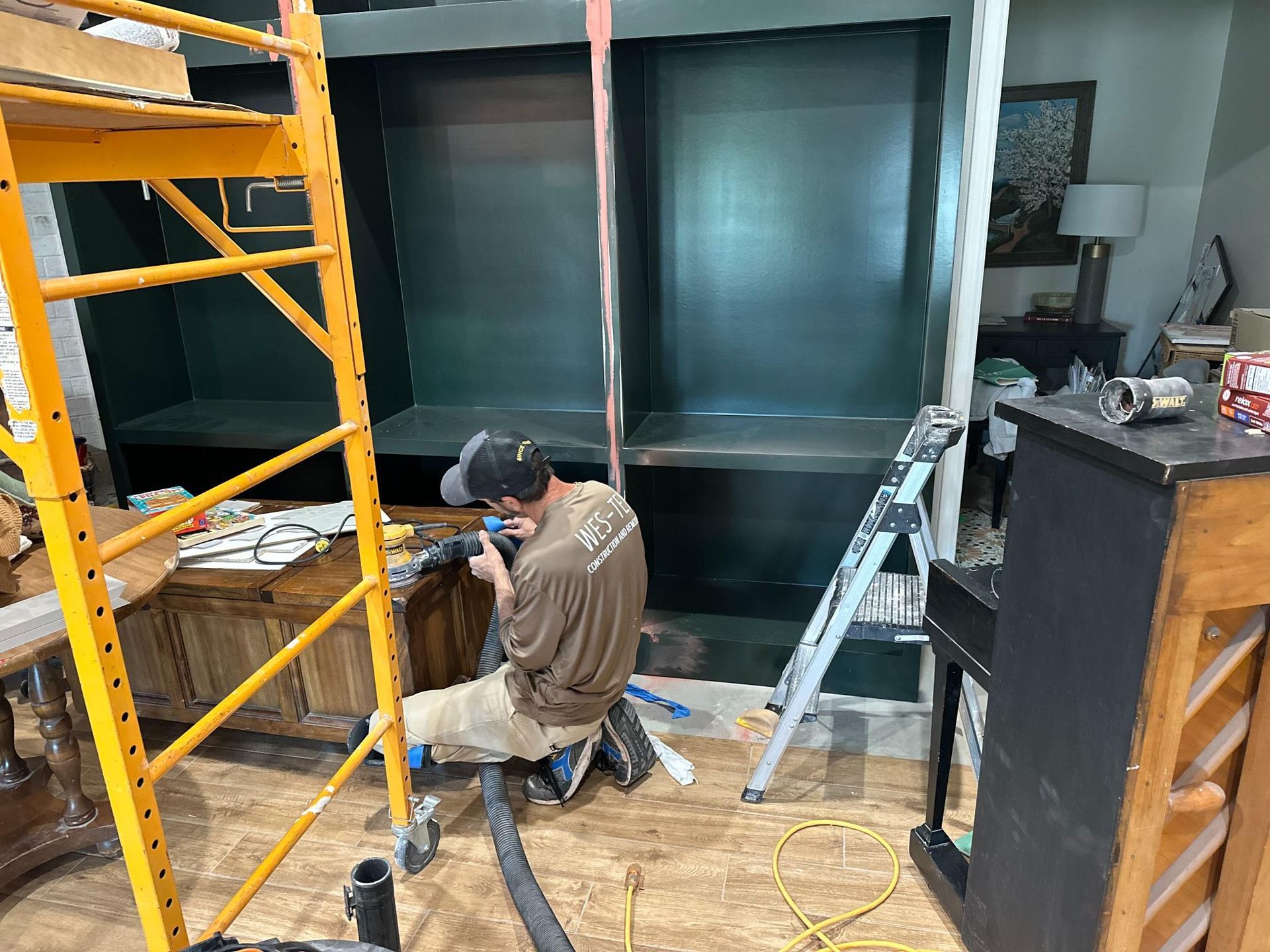 Man installing shelving unit with tools, standing on flooring. Scaffolding, and ladder present. Black and green shelves.