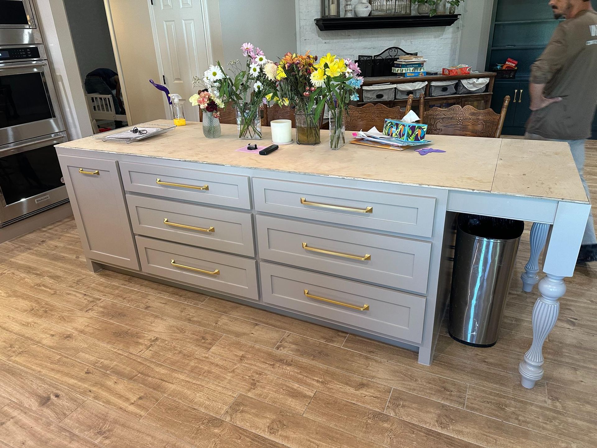 Kitchen island with gray cabinets, gold hardware, and a wood countertop. Flowers and trash can visible.