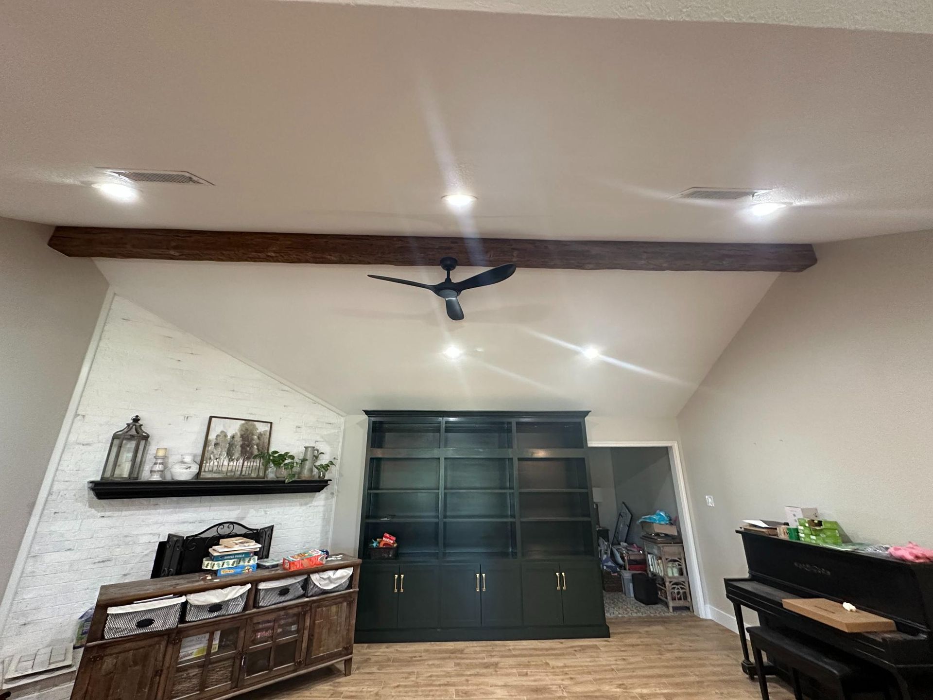 Living room with built-in bookshelves, a brick wall, and a piano. A dark ceiling fan and wooden beam are also present.
