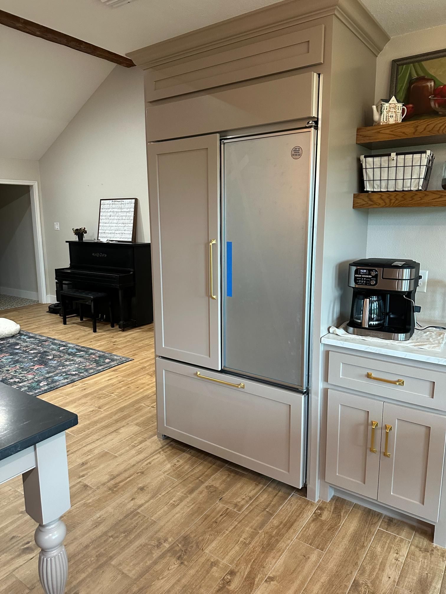 A large refrigerator built into a kitchen cabinet, next to a coffee maker. Light brown cabinets and flooring.