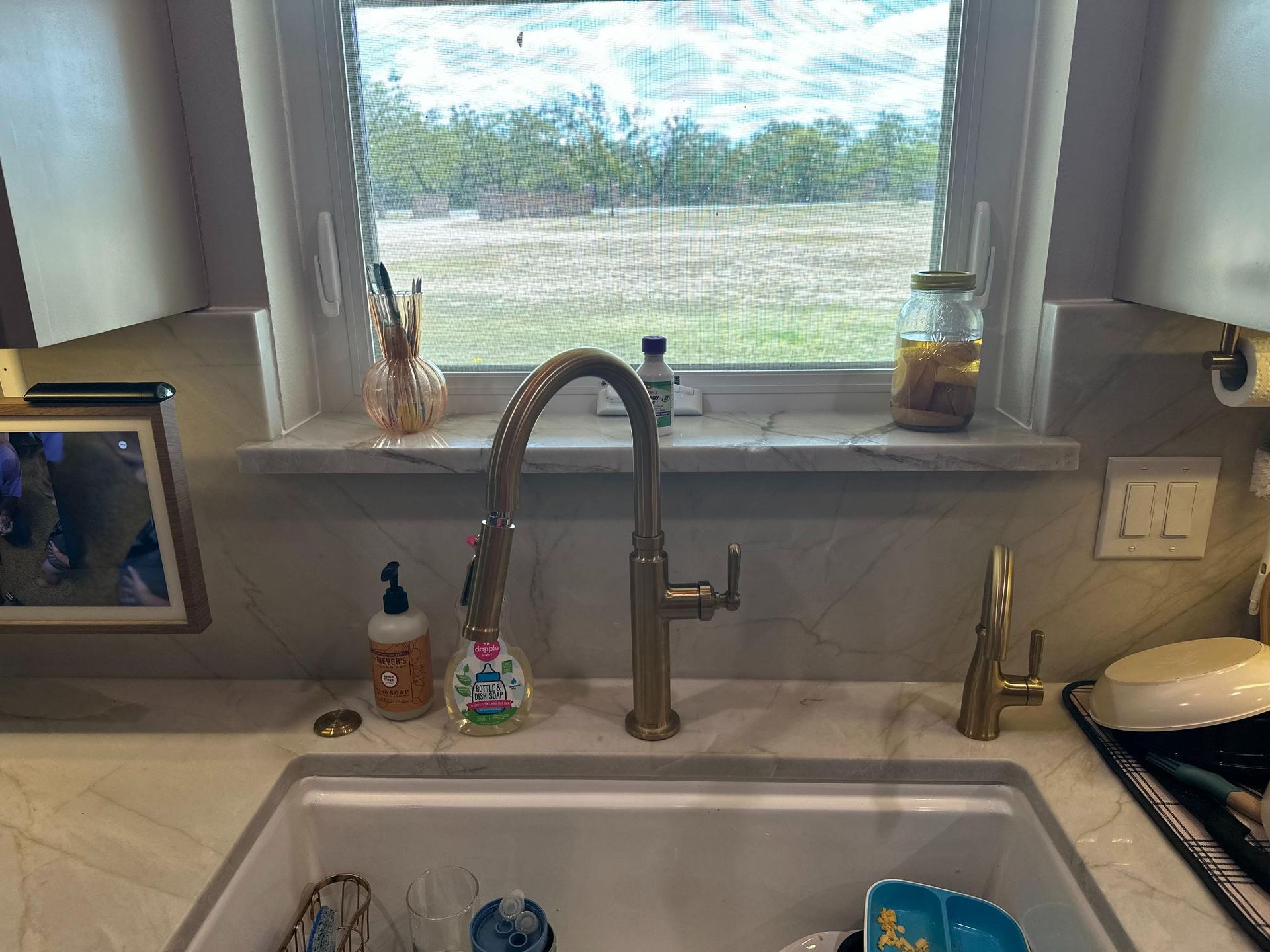 Kitchen sink with gold faucet, window view of field, soap dispenser, and dishes.