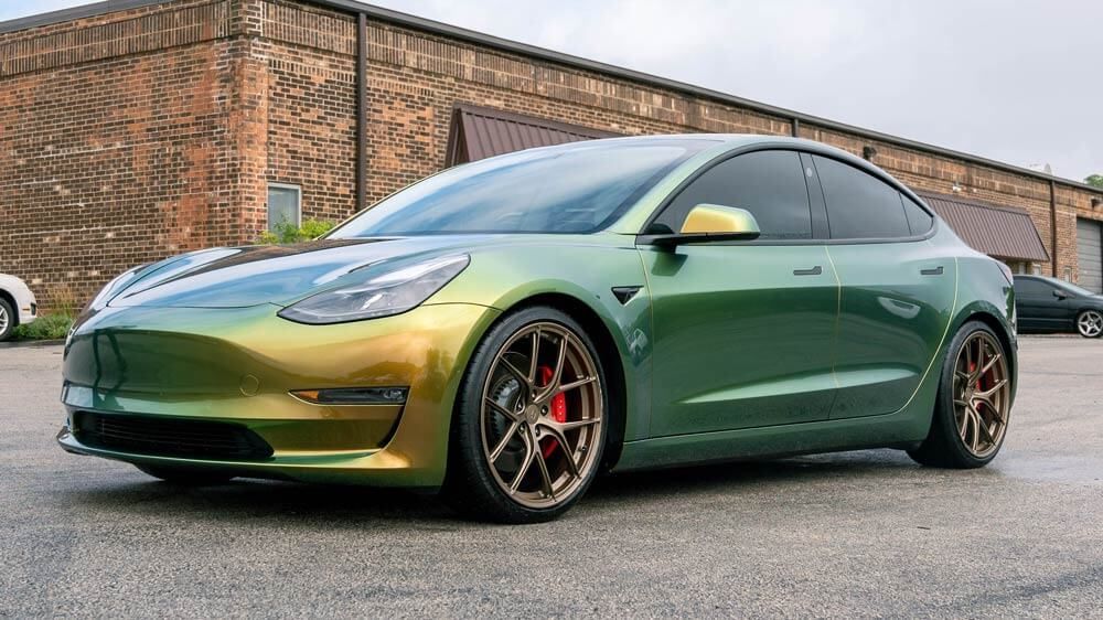 A green tesla model 3 is parked in front of a building.