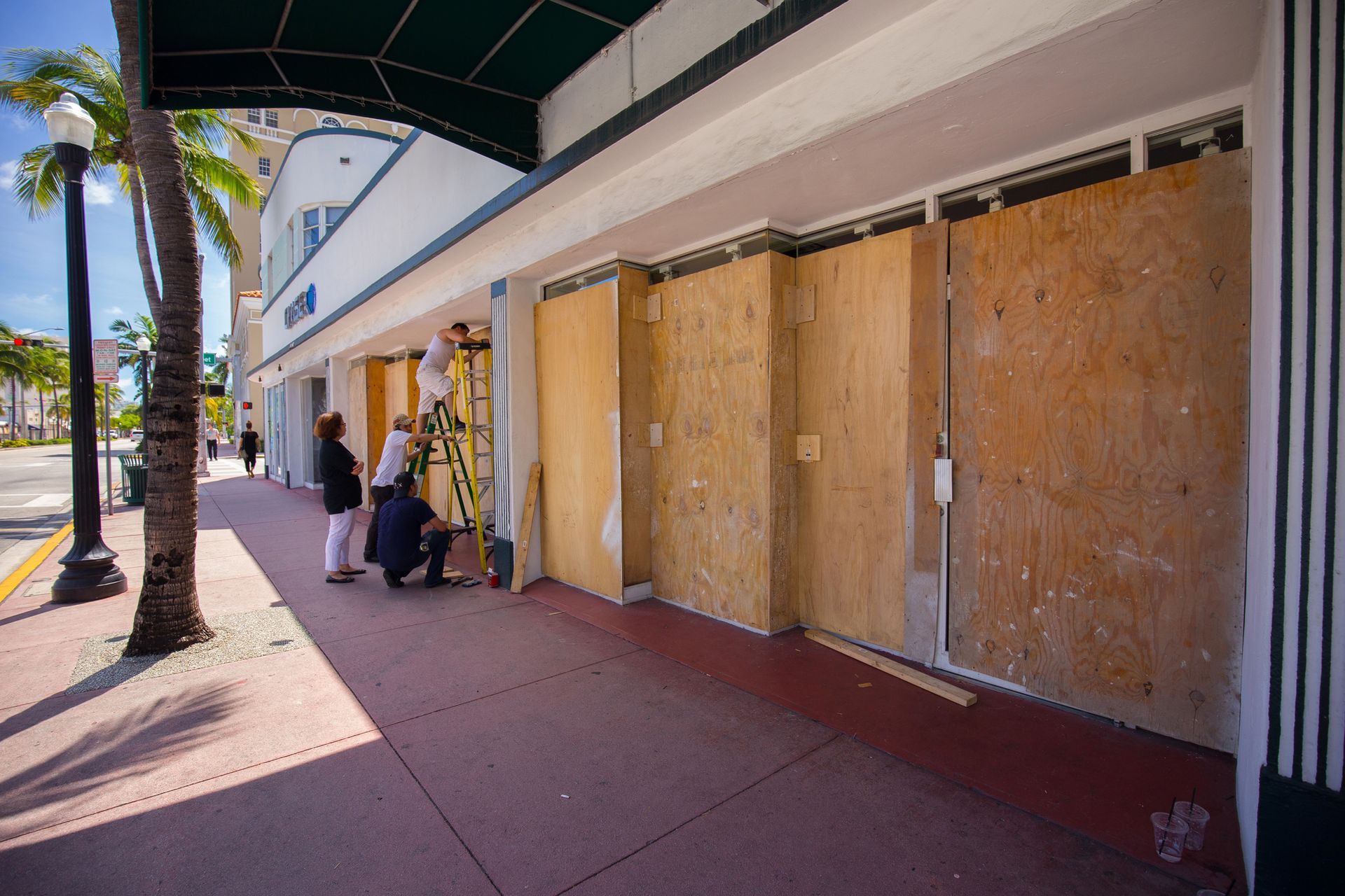A group of people are standing on a sidewalk in front of a building that has been boarded up.