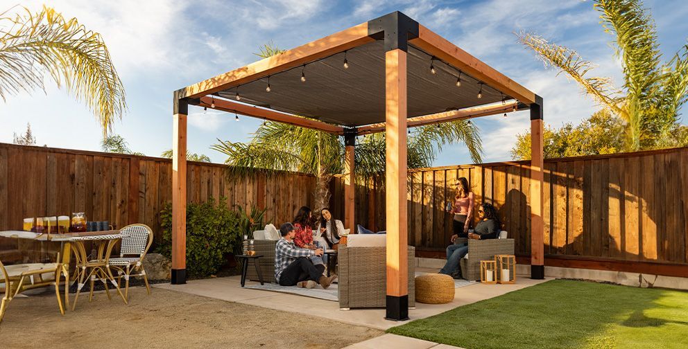 A group of friends under a wooden pergola in a backyard, enjoying drinks on a sunny day.