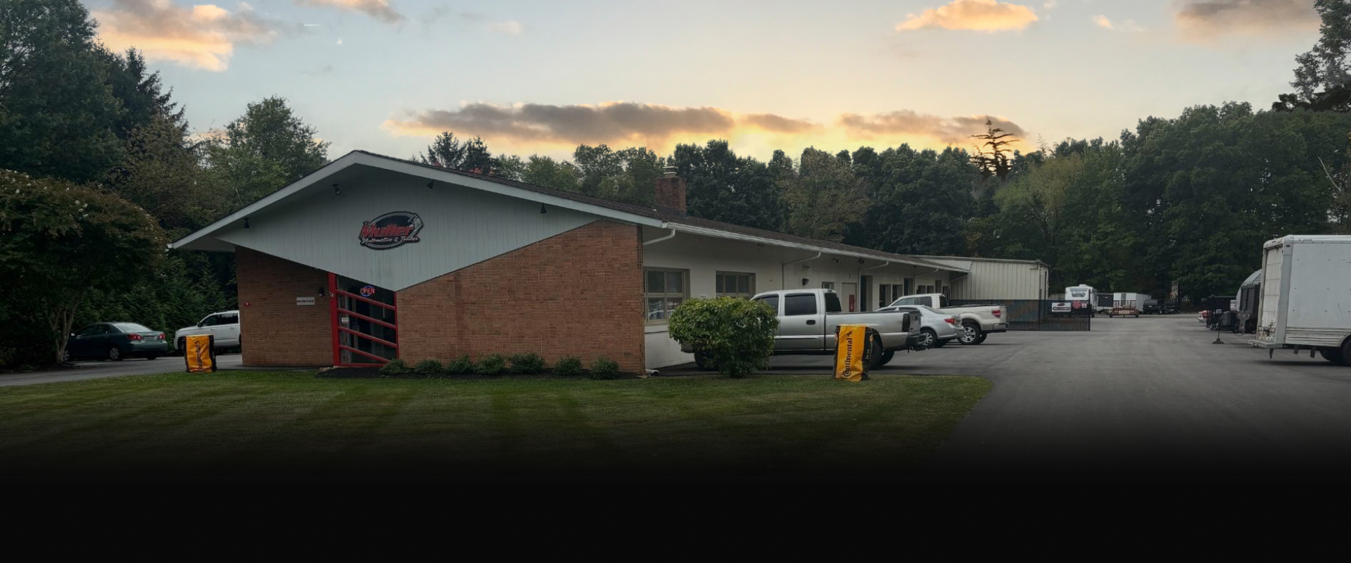 A long brick building with a sloped roof at sunset. Cars and a truck are parked outside. | Dave Muller Automotive & Trailer