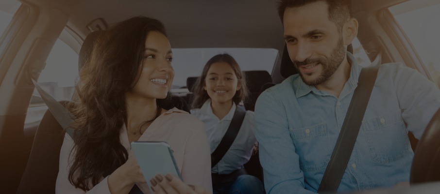 A family of three smiling in a car, woman holding a phone, all wearing seatbelts. | Dave Muller Automotive & Trailer