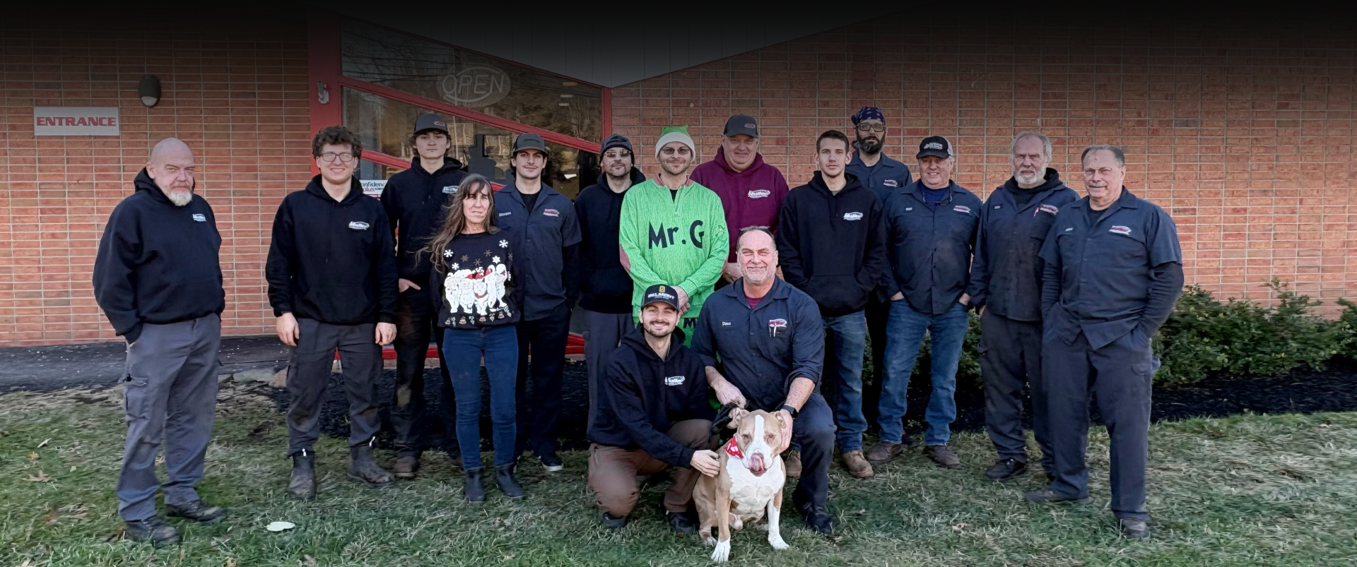 Group of people in black shirts pose outside a brick building, a dog sits in front.