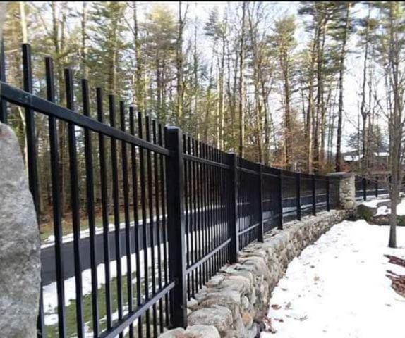 Black metal fence atop a stone wall along a snowy driveway, woods in the background.