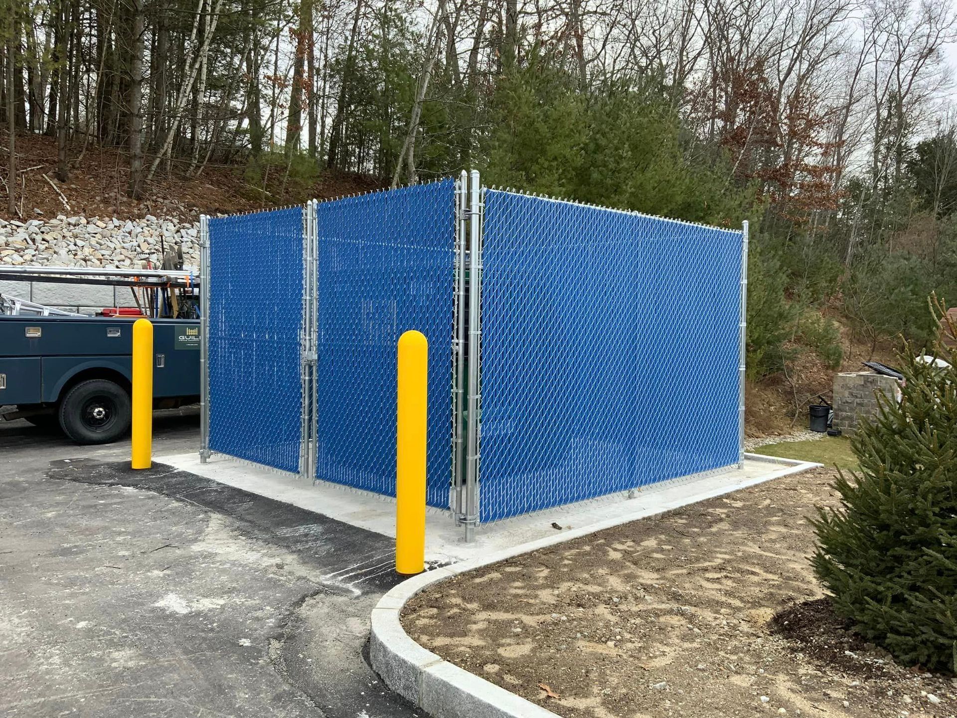 Blue screened enclosure, possibly for utilities, with yellow bollards in a parking area, trees in the background.