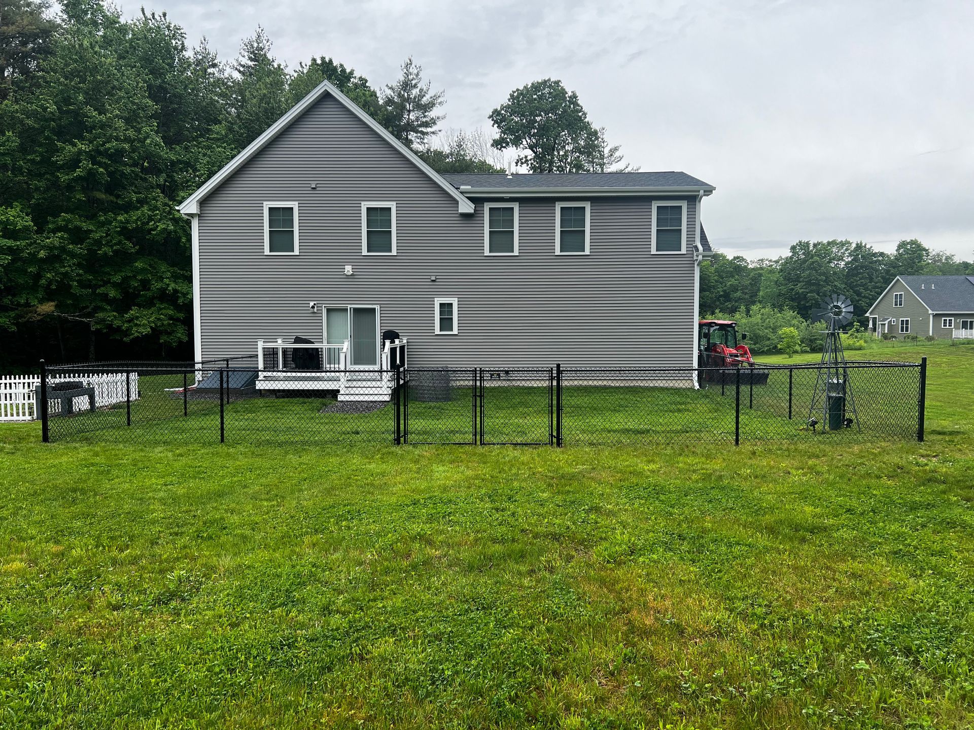 Gray house with black chain-link fence on grassy lot; trees in background; cloudy sky.
