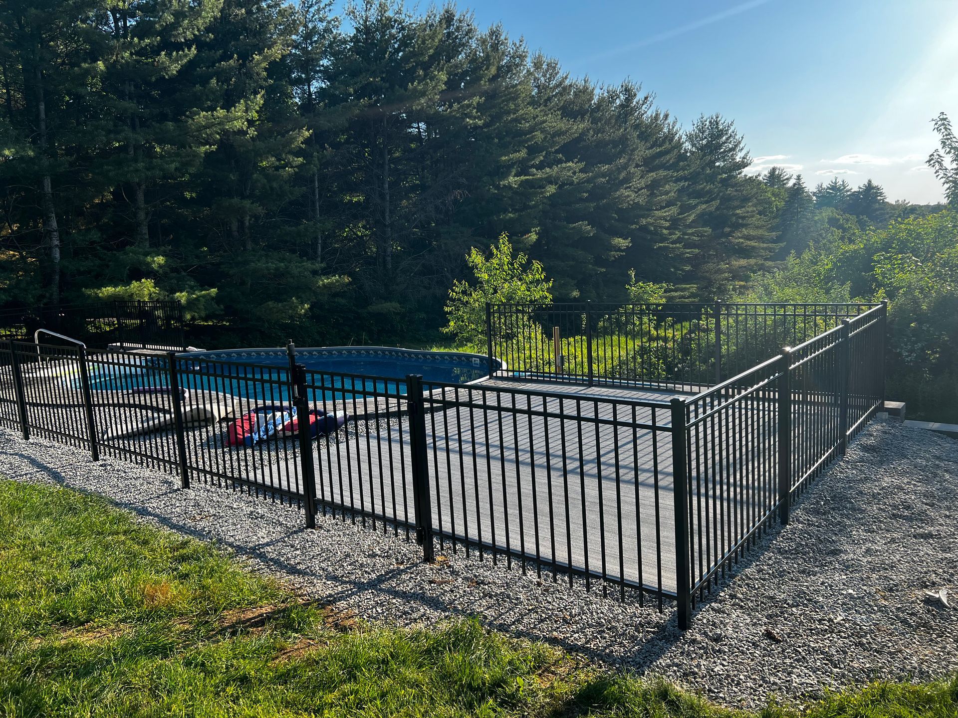 Pool surrounded by a black fence. Gravel base with lush green grass and trees in the background.