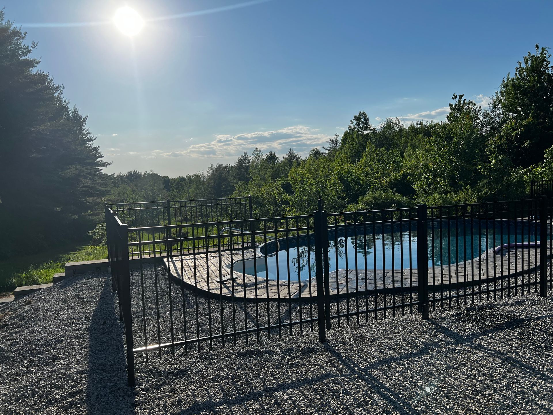 A pool enclosed by a black wrought iron fence, with a sunny sky and trees in the background.