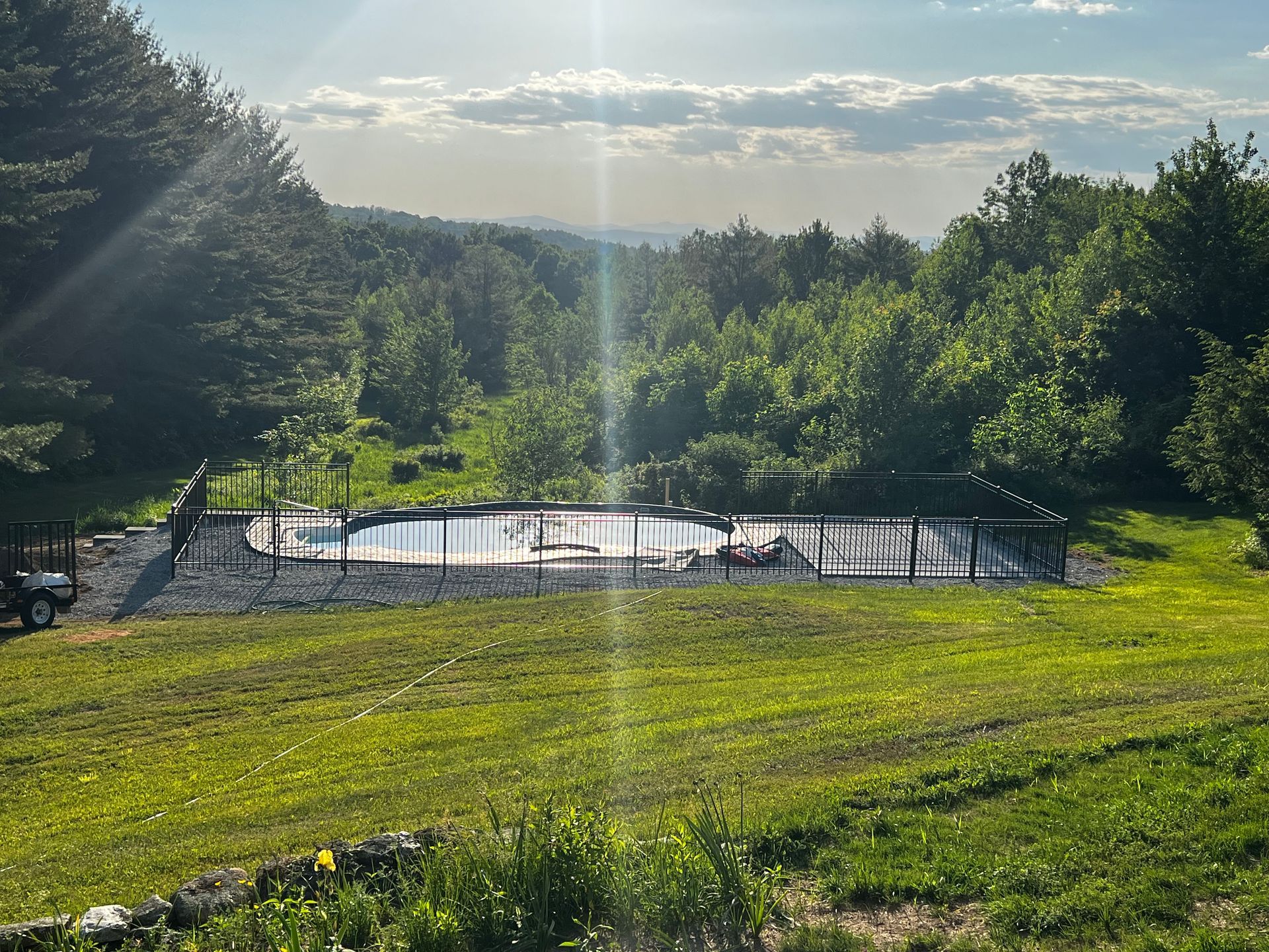 A partially constructed outdoor swimming pool surrounded by green grass and trees, under a sunny sky.