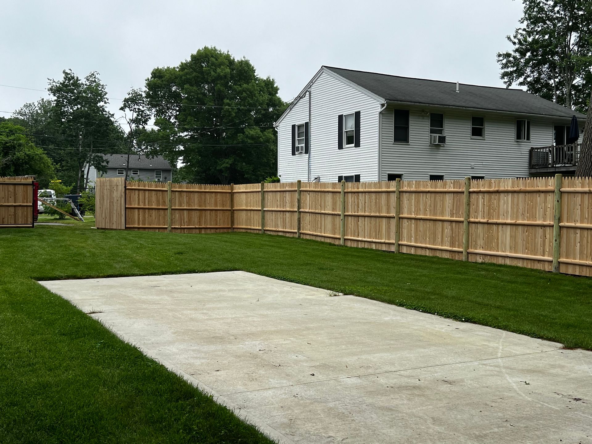 Backyard with concrete pad and wooden fence, next to a multi-story house and shed. Green grass surrounds.