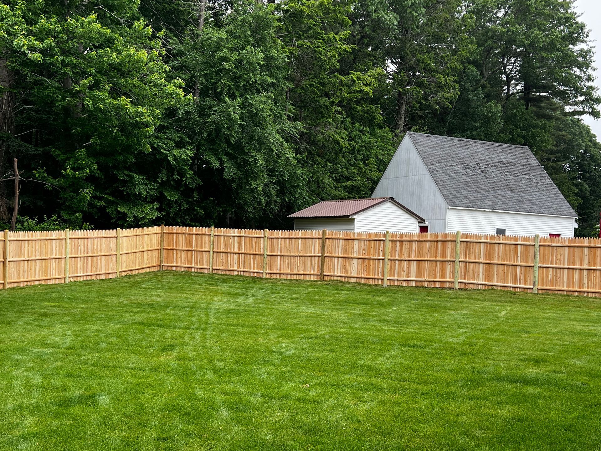 A new wooden fence surrounds a green lawn in front of a house and trees.