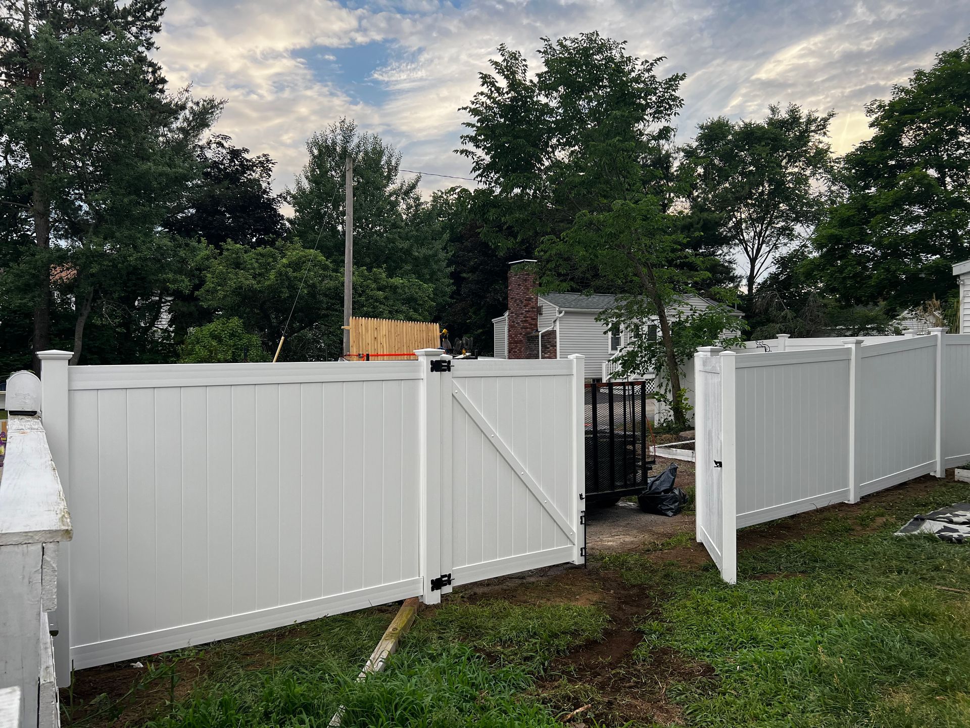 White vinyl fence with gate in a yard with grass and trees under a cloudy sky.