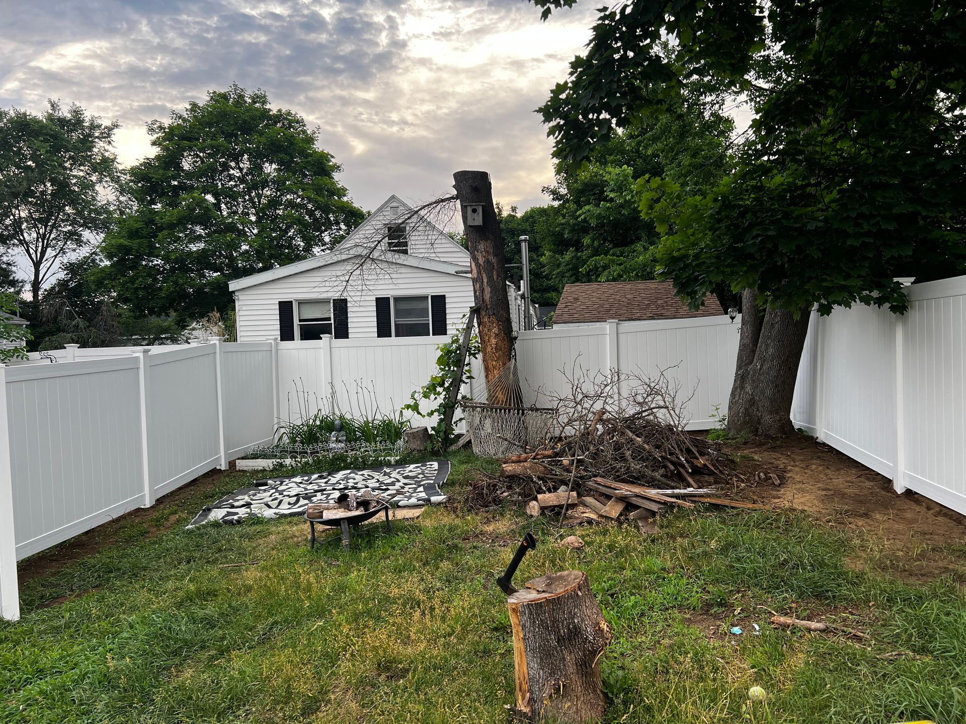 Backyard with a cut tree, debris, white fence, and small house. Overcast sky.