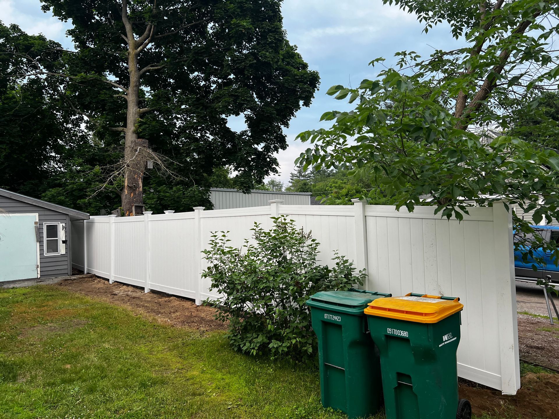 White vinyl fence in a backyard setting with green trash bins.