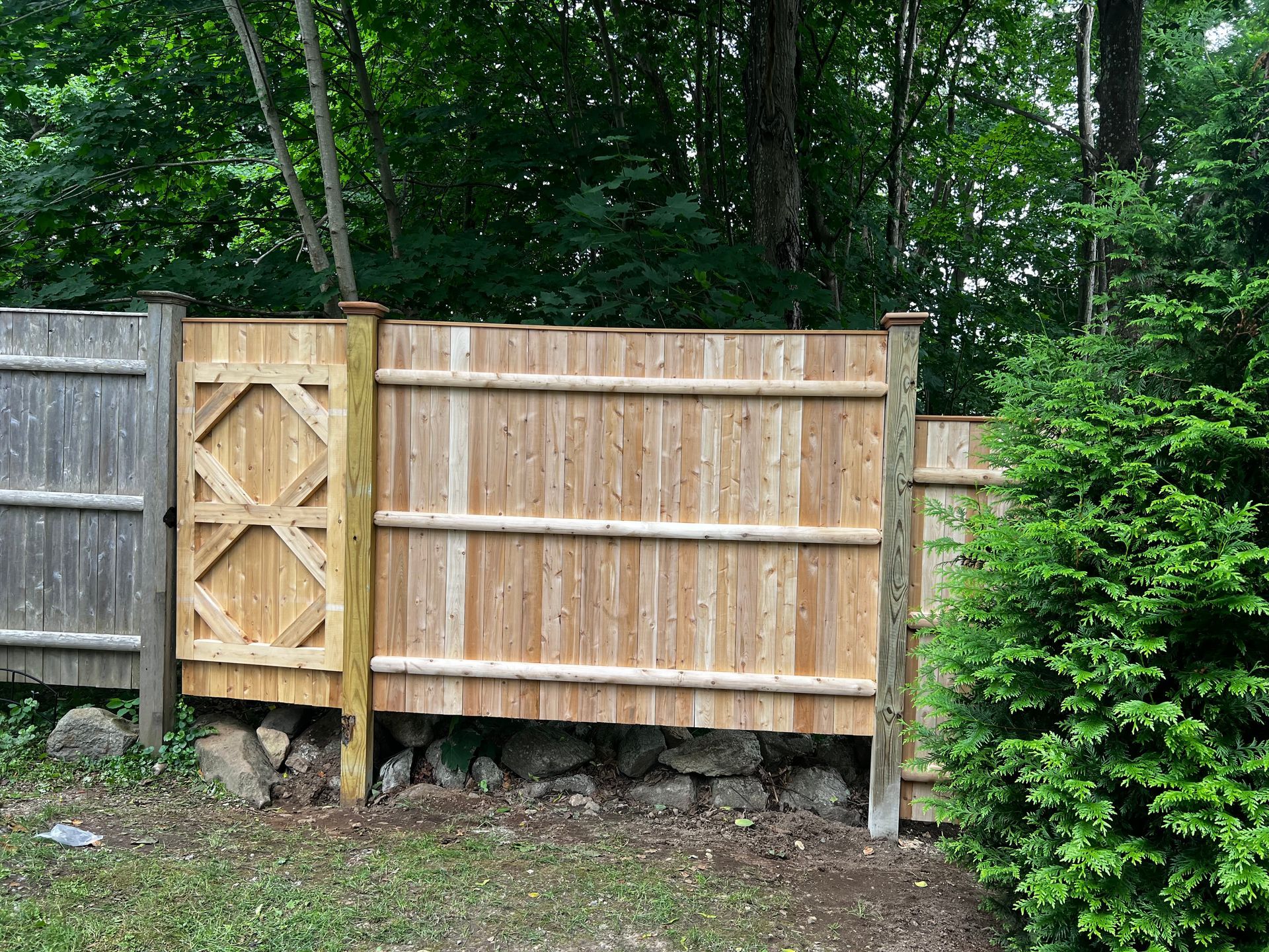 Wooden fence section with gate and rock base, surrounded by trees.