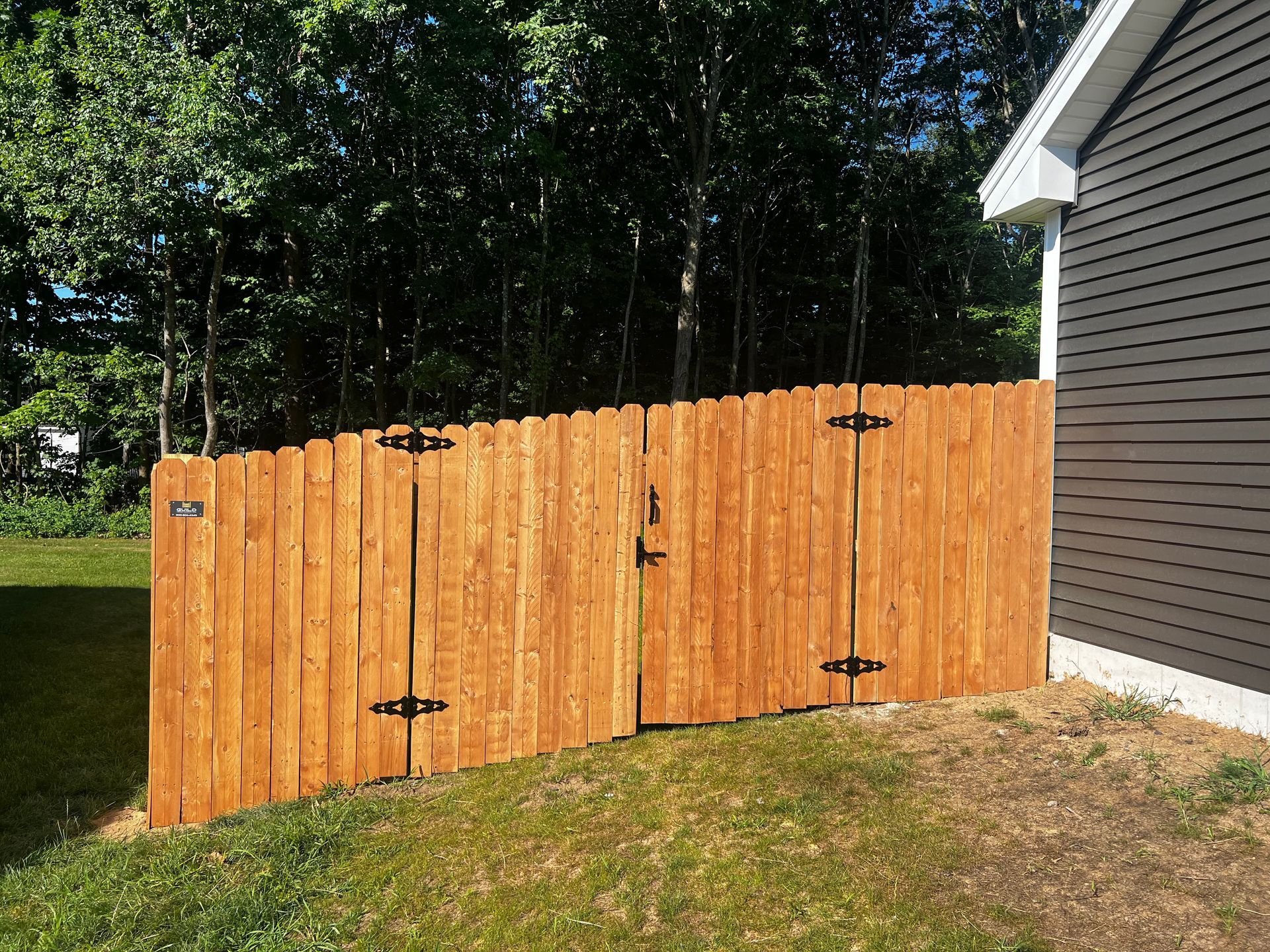 Wooden fence with gate, next to a house, brown, green grass, trees in the background.