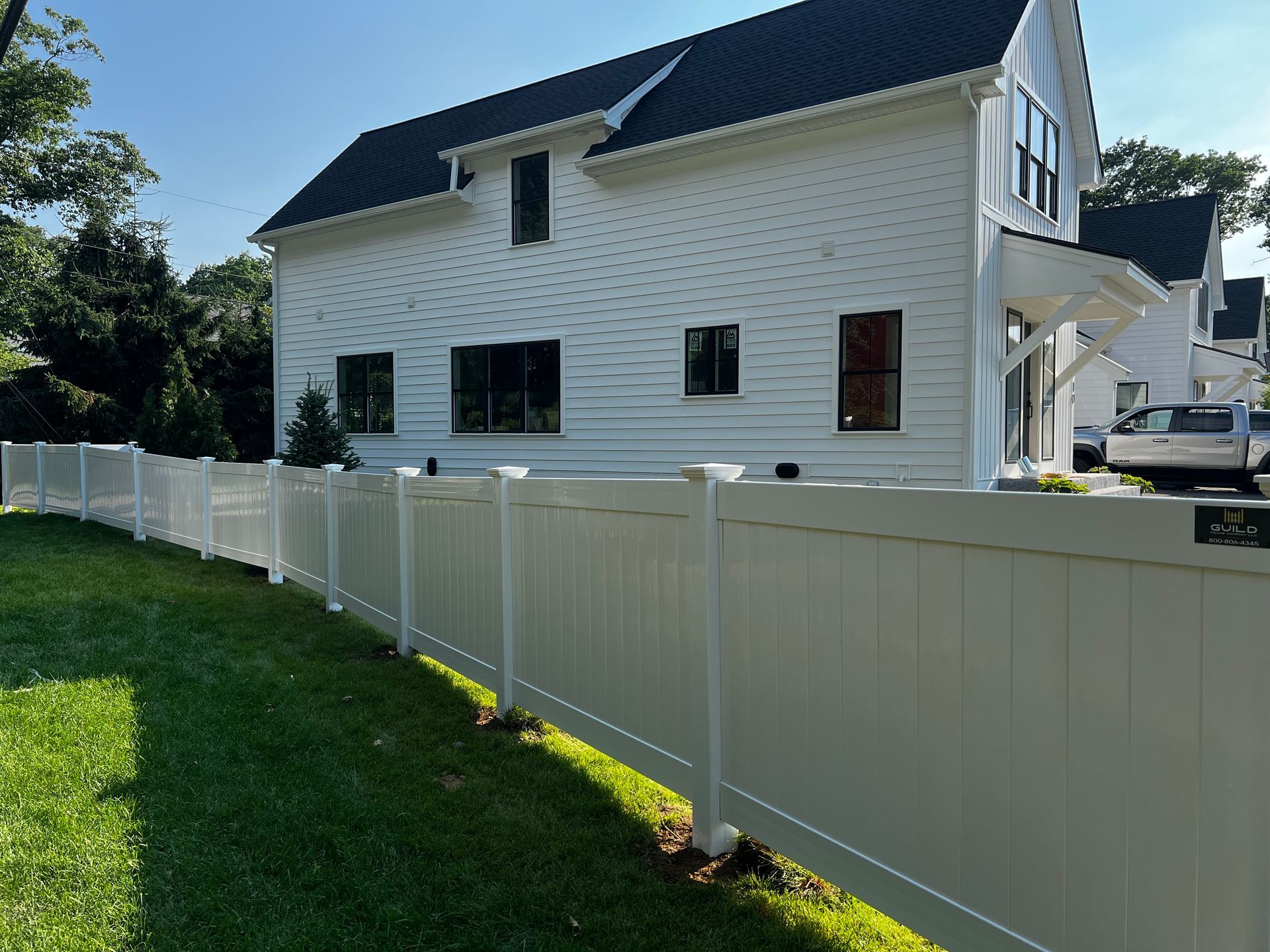 White vinyl fence surrounds a white house with black roof, on green grass under a clear blue sky.