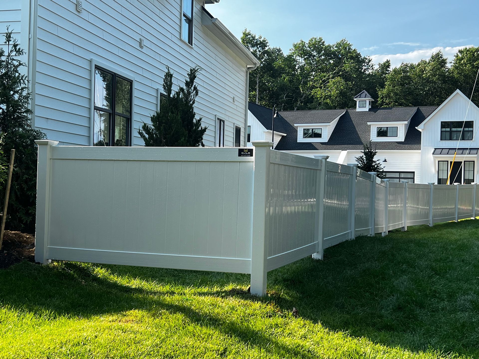 White fence surrounding a lawn, next to a white house with trees in the background.