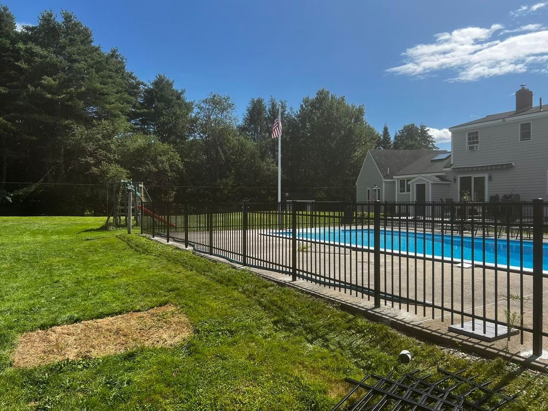 Black fence surrounds a backyard pool. Green grass, trees, and a house are in the background. Blue sky overhead.