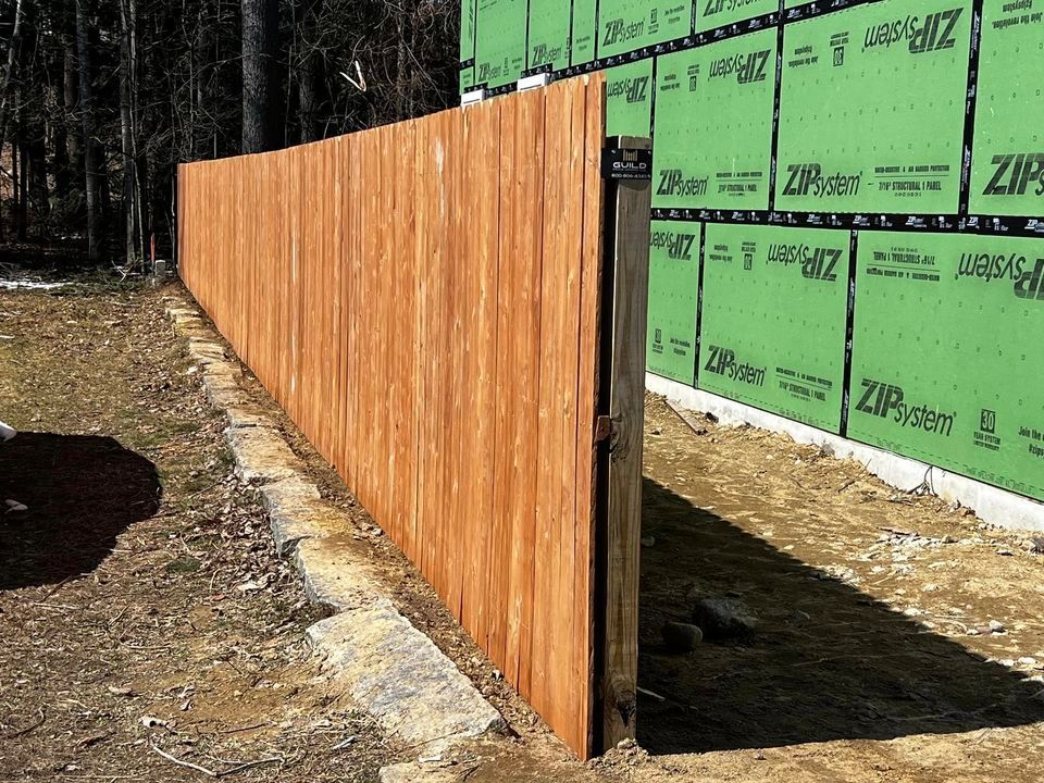 Wooden fence, stained brown, next to a building with green siding. Sunny outdoor setting.