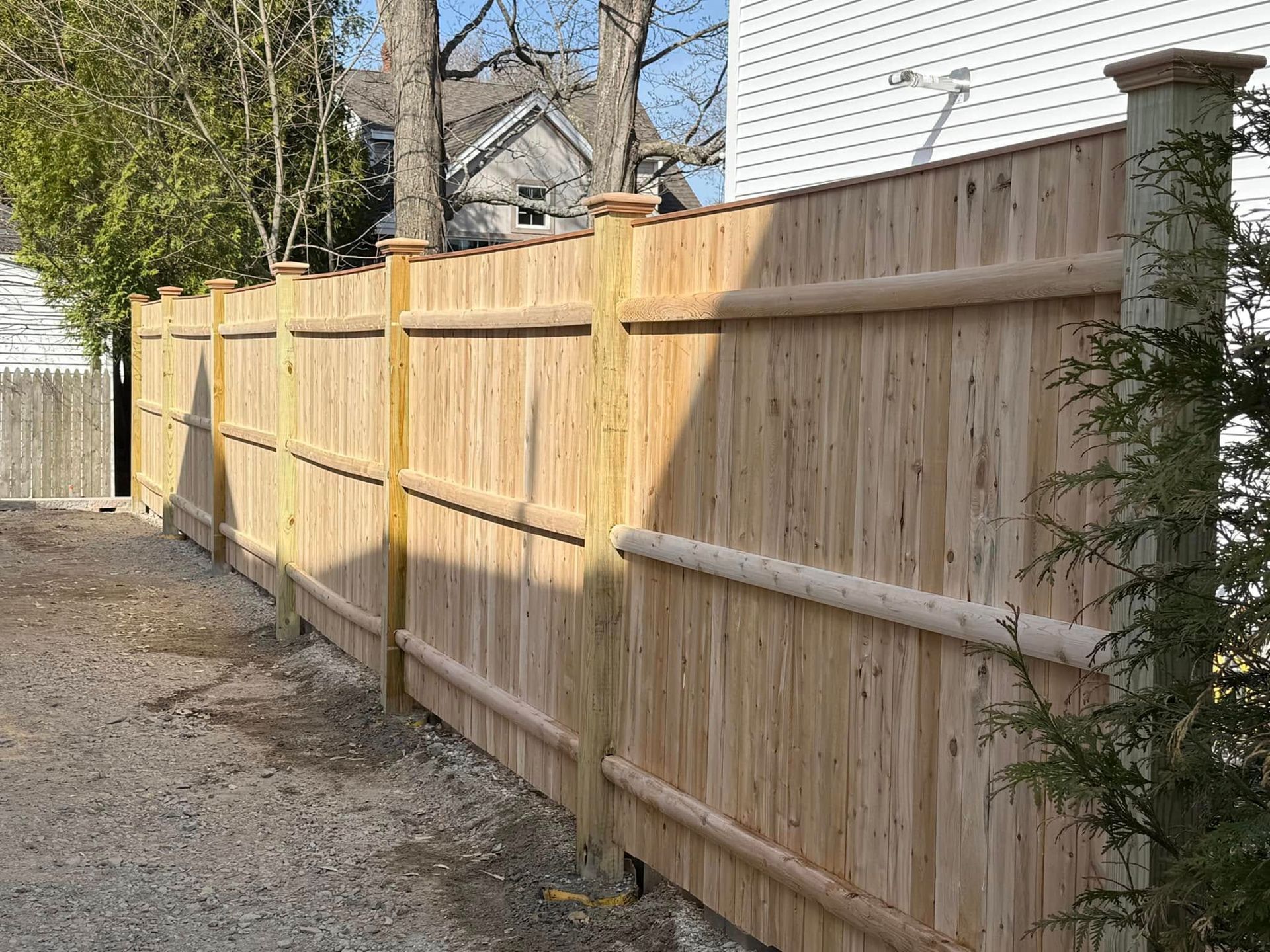 Wooden fence along a gravel path with a house in the background and a tree on either side.
