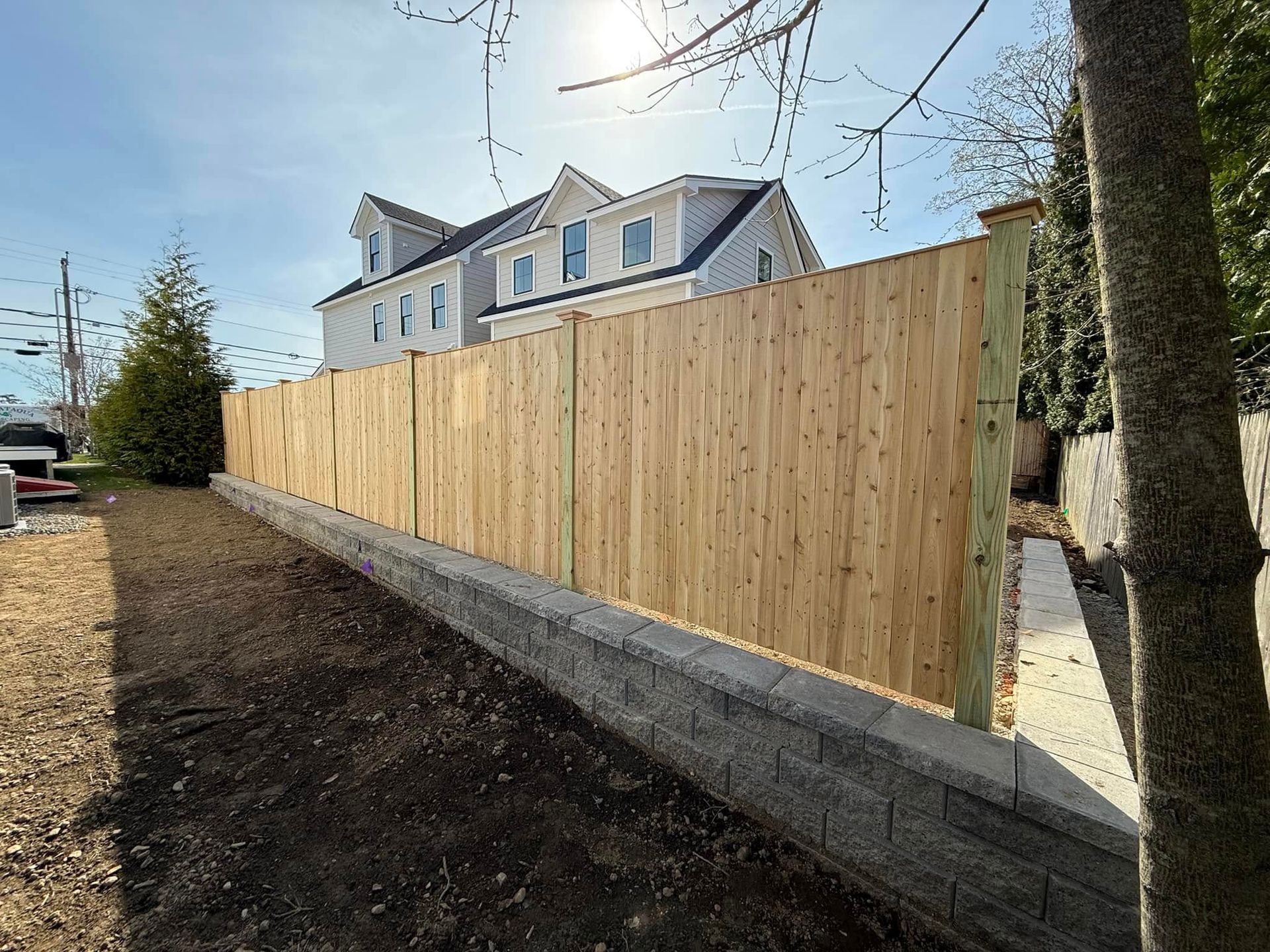 Wooden fence atop a retaining wall, with a light-colored house in the background under a blue sky.