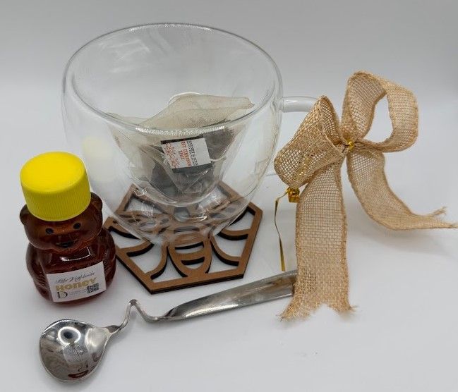 Heart-shaped glass tea cup holding honey bear and tea bags, displayed on wooden bee coaster with bow.