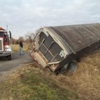 A semi-truck trailer tipped over on its side next to a road, with a tow truck nearby.