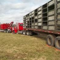 Red semi-truck towing trailer loaded with stacked gray metal structures on a grassy field under a cloudy sky.