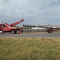A red tow truck hauling a long auger on a trailer, on a road with grass.