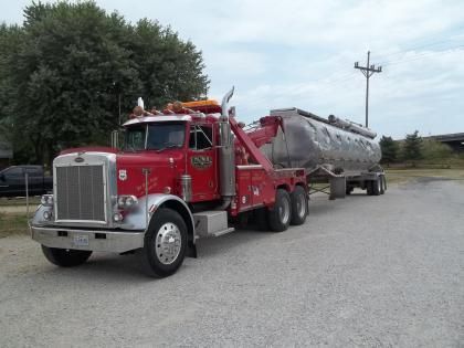 Red Peterbilt tow truck towing a silver tanker trailer on a gravel lot under a cloudy sky.