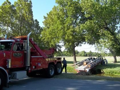 Red tow truck pulling a trailer from a grassy ditch near trees. A person stands nearby.