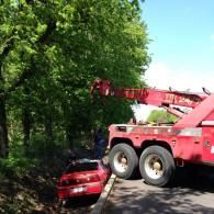 A red tow truck lifting a red car out of a ditch next to trees.