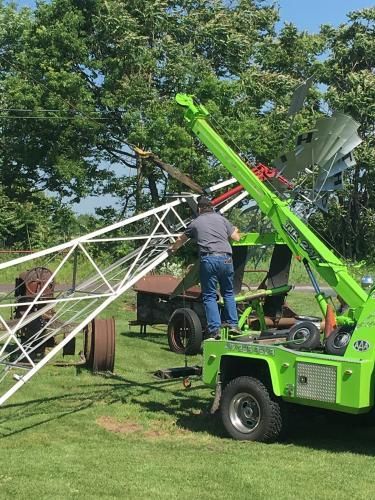 Man using a green crane to lower a metal tower section in a grassy area with trees.
