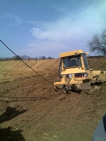 Yellow bulldozer stuck in muddy field, cable pulling, blue sky.