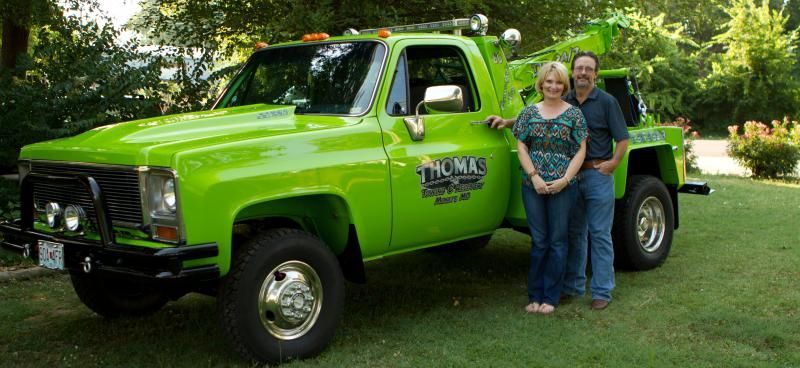 A green tow truck with two people posing next to it. They are in a grassy setting.