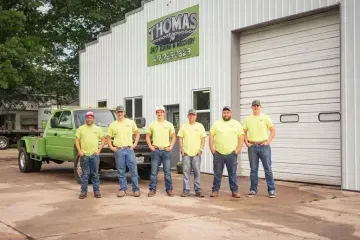 Six men in yellow shirts and jeans stand in front of Thomas Dirt Work & Services building with a green truck.