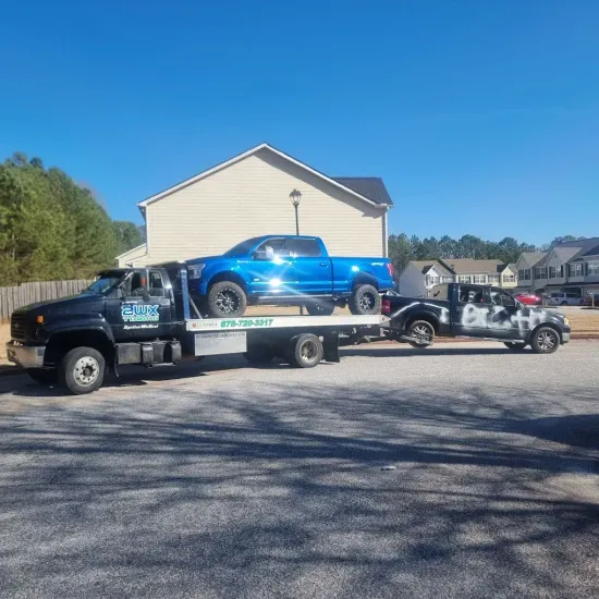 A blue truck on a tow truck, which is towing a black and white truck, parked on a paved road.