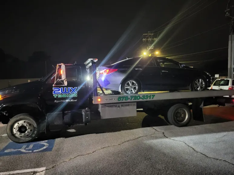 Black tow truck hauling a dark car at night. The truck is parked near a curb.