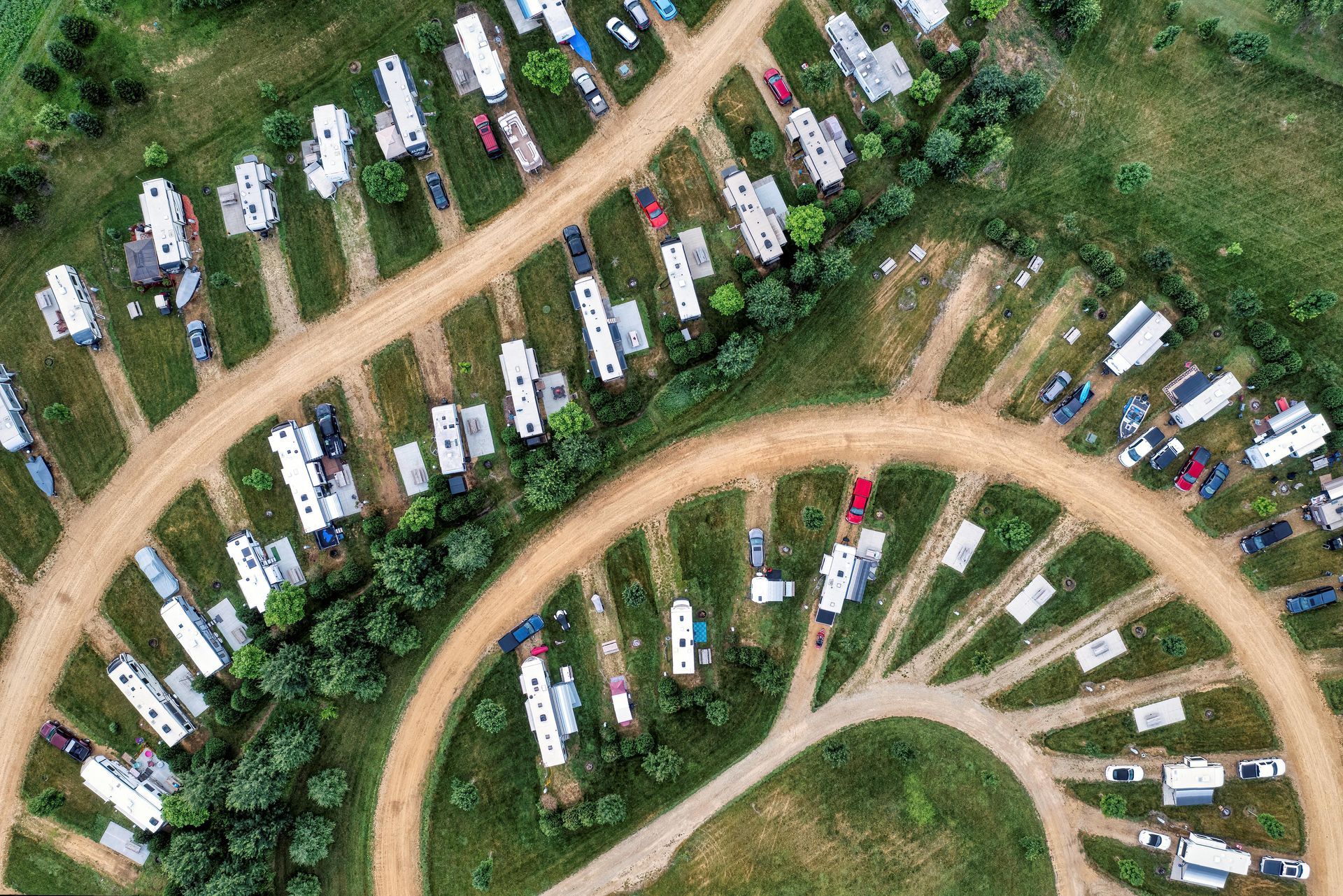 Aerial view of rows of white RVs and campers parked along curved dirt roads in a green campground.