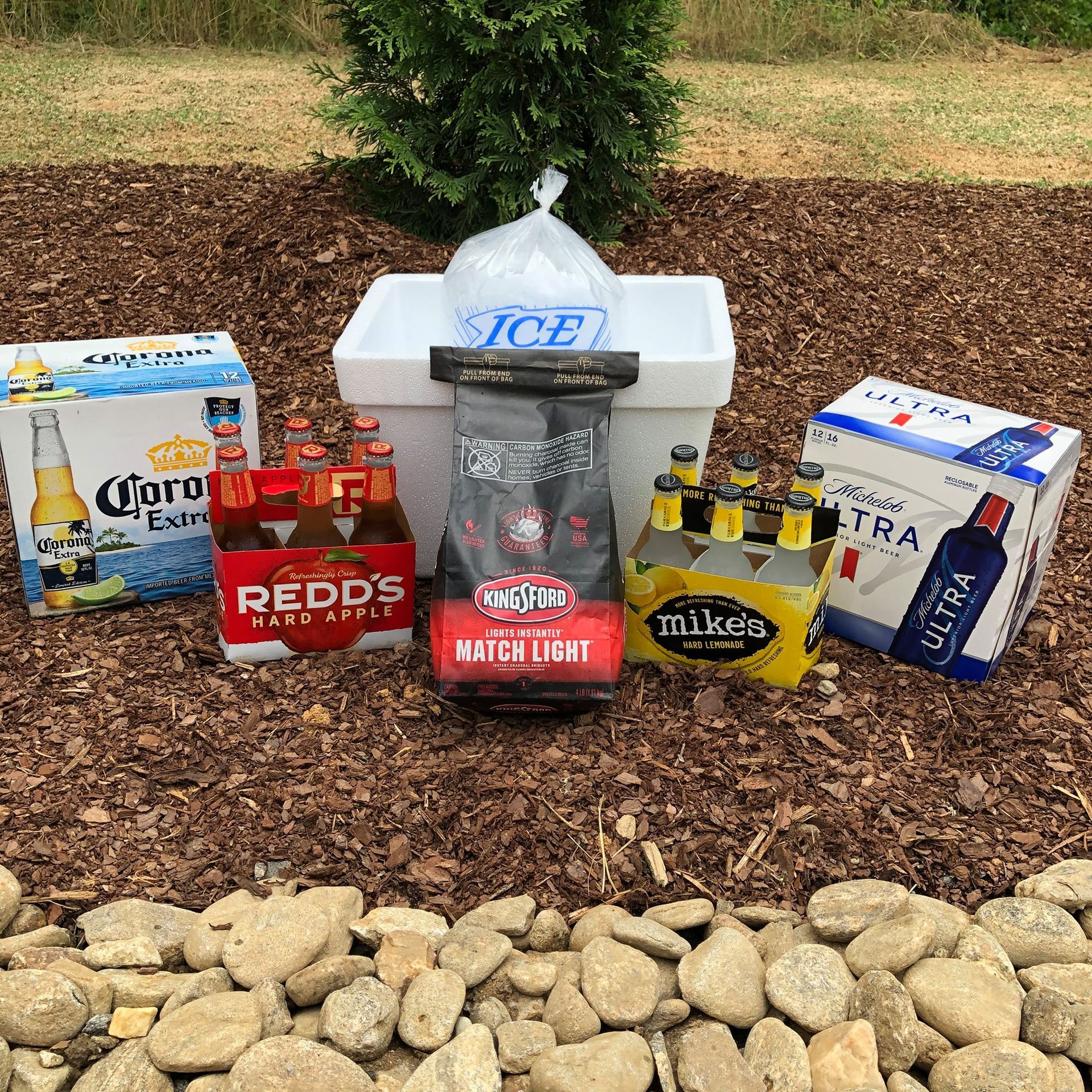 A cooler with ice, charcoal, and beer: Corona, Redd's, and Michelob Ultra, on wood chips, near rocks.