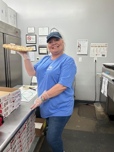 A smiling person in a blue shirt and cap stands in a kitchen, holding a pizza on a tray near stacks of pizza boxes.