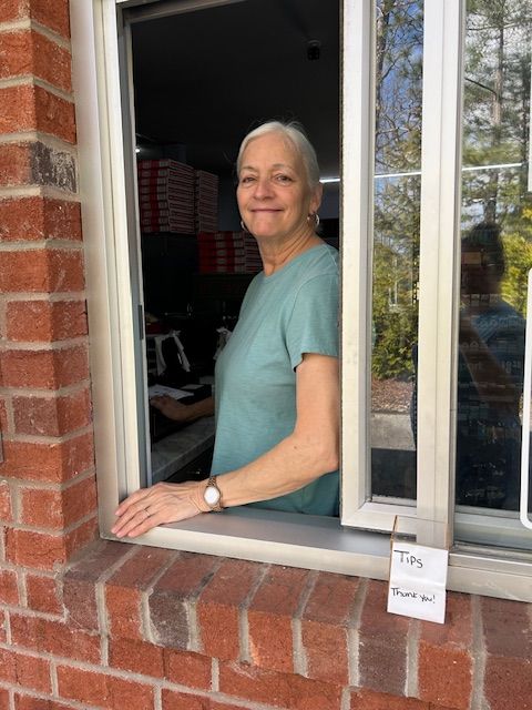 A person wearing a teal t-shirt leans out of a brick building's drive-thru window, smiling at the camera.